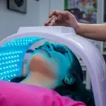 Unrecognizable woman with dark hair with pierced ears lying under ultraviolet lamp while visiting contemporary clinic and spending time in modern salon